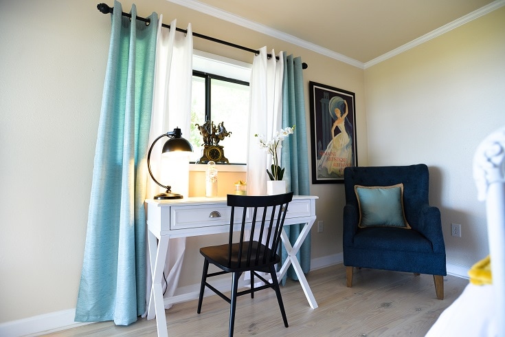 A bright workspace features a white desk illuminated by a stylish lamp. A black chair is positioned in front of the desk. Natural light enters through a window dressed with teal and white curtains. A cozy armchair adds a touch of comfort to the corner.