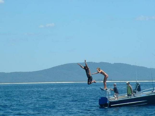 A group of individuals is seen from a distance enjoying a day on the water, with two people leaping off the side of a boat into the calm blue lake. A scenic mountain range can be observed in the background under a clear sky.