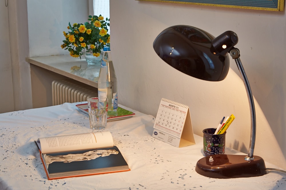 A desk is set with a study lamp, a glass of water, and an open book. A calendar and a cup containing pens are also present. A vase of yellow flowers adds a touch of color to the bright workspace.