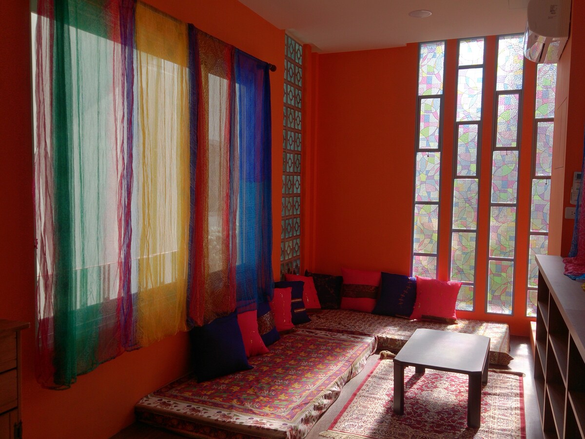 A lively seating area features colorful fabric drapes and vibrant walls. Cushions in various shades provide comfortable seating around a low table. Stained glass windows add an artistic touch, allowing natural light to filter in, creating a warm ambiance.