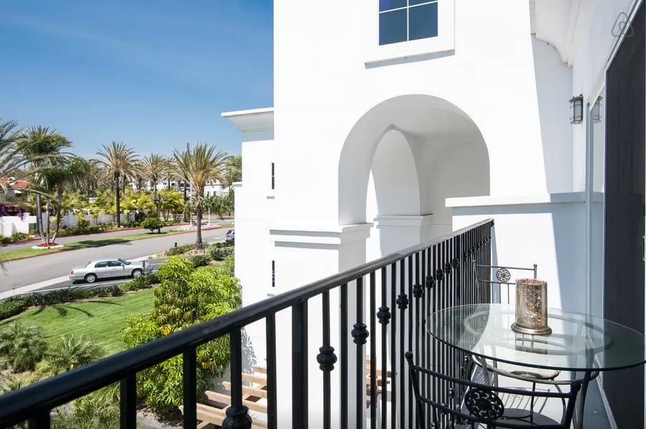 A private balcony is visible, featuring a round glass table with a candle. The black railing frames the view of the surrounding greenery and palm trees, while a pathway leads to the street below, enhancing the sense of openness.