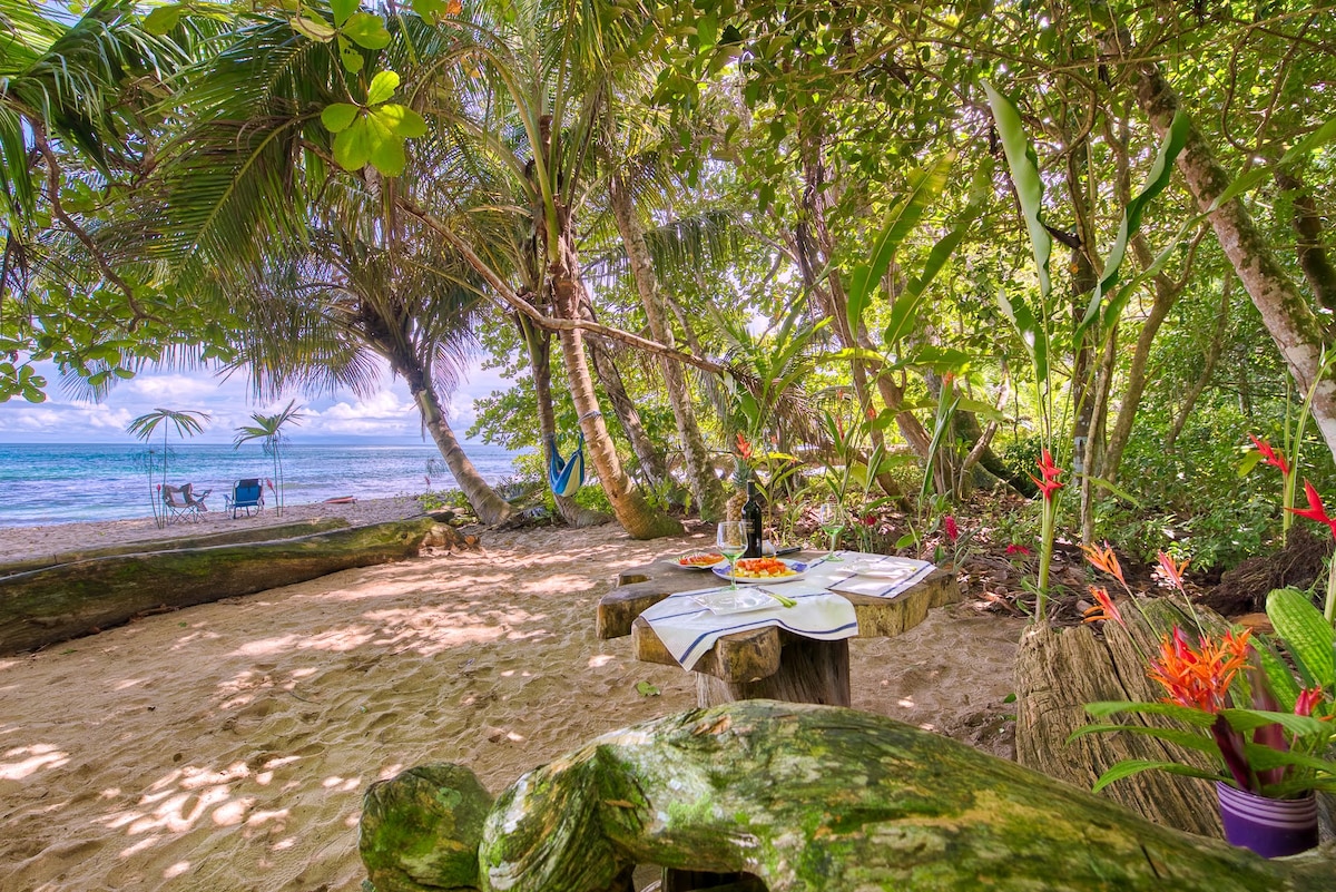 A shaded dining area features a rustic wooden table set with tableware, surrounded by lush tropical greenery. In the background, a view of the calm ocean can be seen through the trees, alongside a few beach chairs for relaxation.