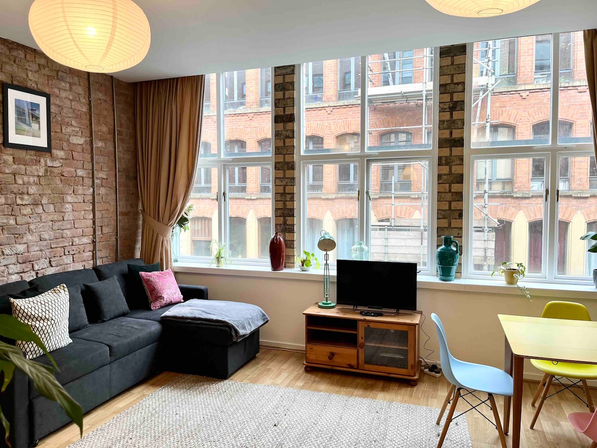 An open plan living area features large windows with natural light illuminating the space. Original brick walls highlight the warmth of the room. A dark sofa is decorated with cushions, while a wooden TV stand and a small dining table with colorful chairs complete the ambiance.