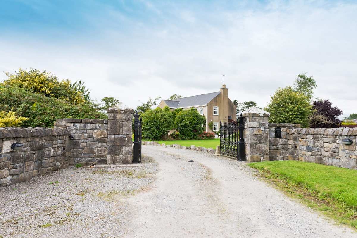 A gravel driveway leads through open gates, framed by low stone walls adorned with greenery. The two-story country home is visible in the background, surrounded by lush gardens and trees under a cloudy sky.