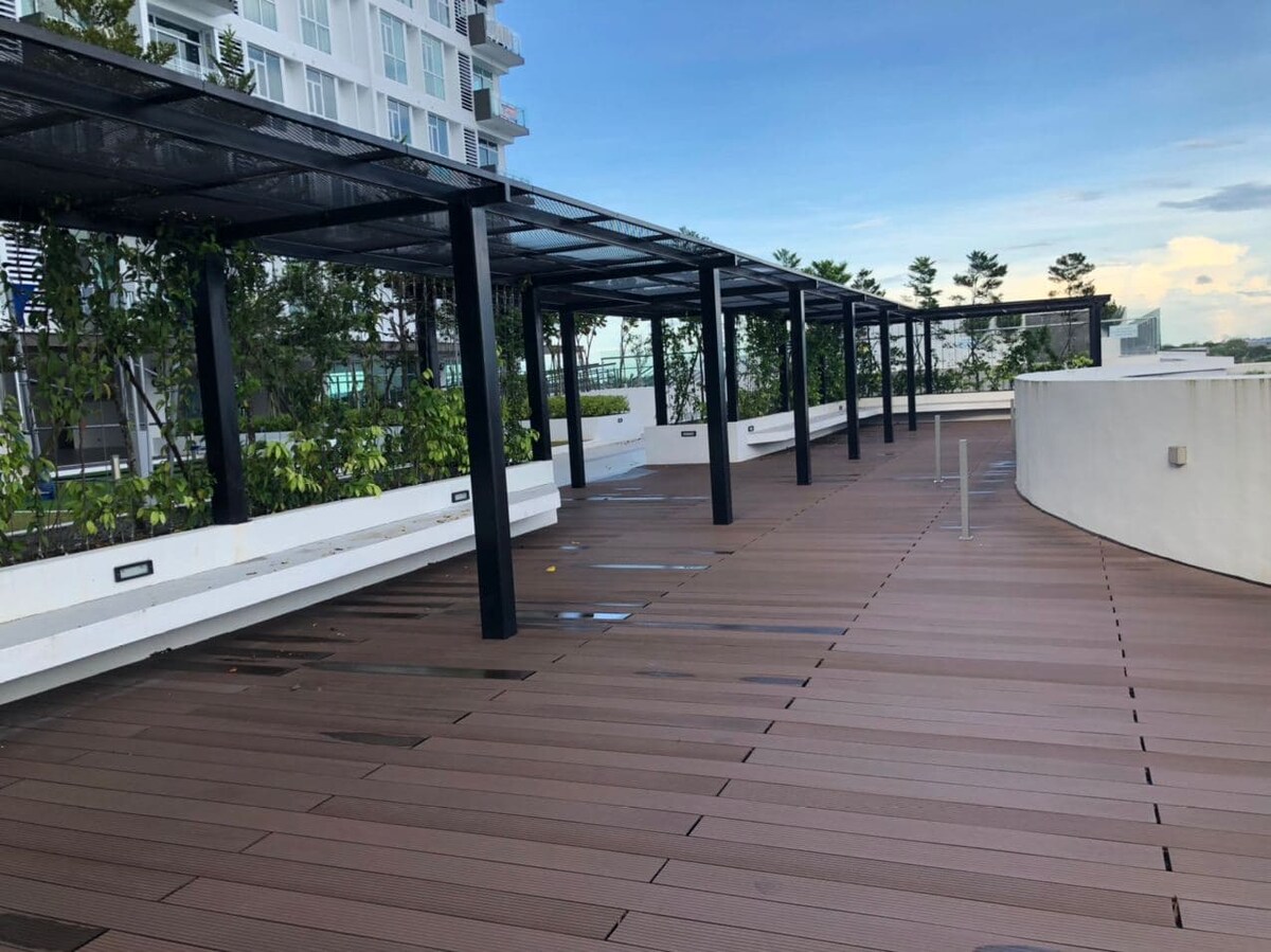 A spacious outdoor area features wooden decking surrounded by green landscaping. Canopy structures provide shade for the seating areas, enhancing comfort and relaxation. The open space is ideal for enjoying fresh air and views of the surroundings, inviting a tranquil retreat.