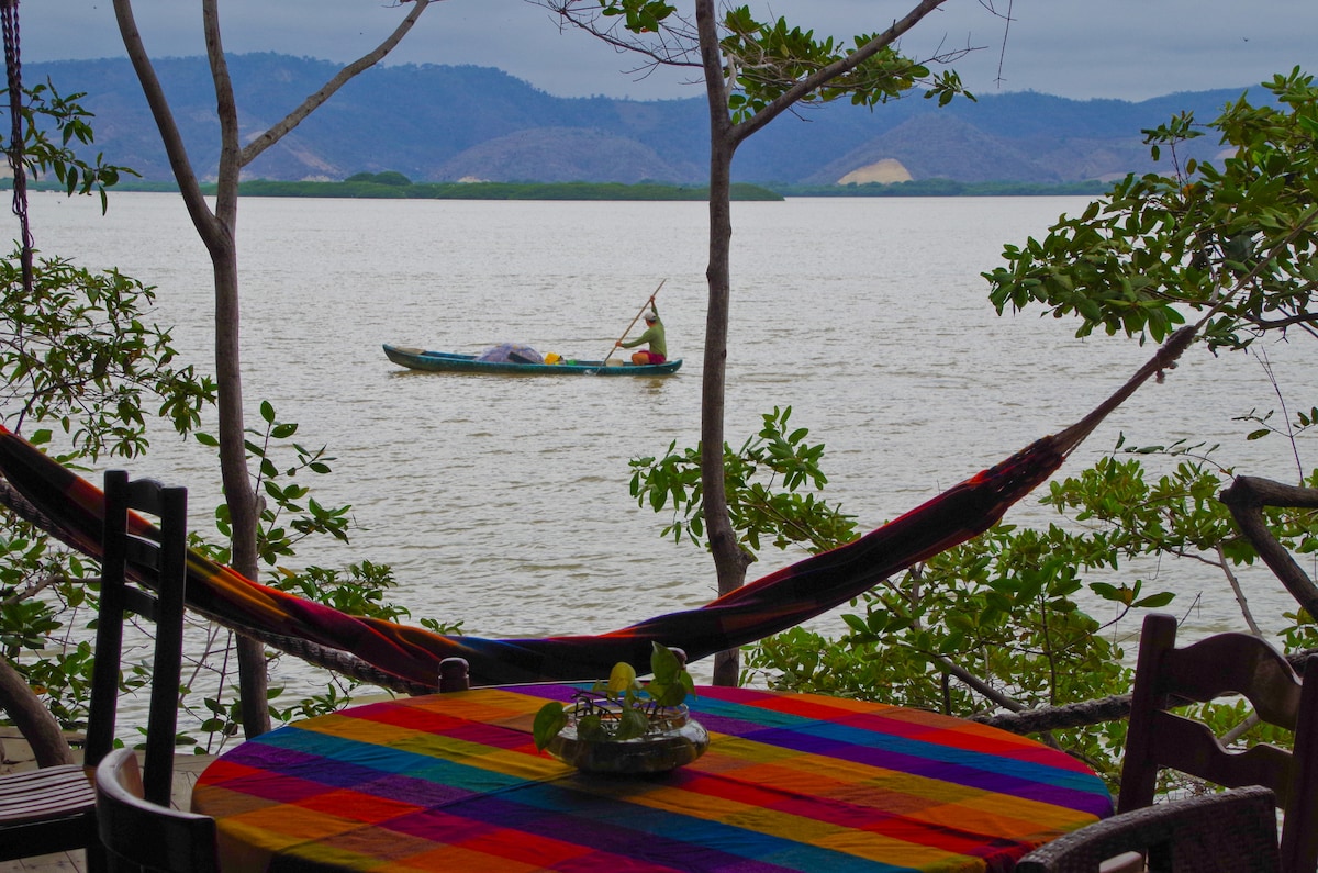 A colorful tablecloth adorns a round table set near the water, framed by lush greenery and tree branches. A hammock is visible beside the table. In the distance, a small boat glides across the calm water, against a backdrop of rolling hills and a cloudy sky.