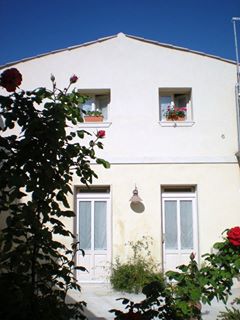The image features a charming building exterior with two white-paneled doors and two small windows above, adorned with potted plants. The soft yellow facade is framed by blooming rose bushes, adding a natural touch to the entrance. A clear blue sky provides a bright backdrop.