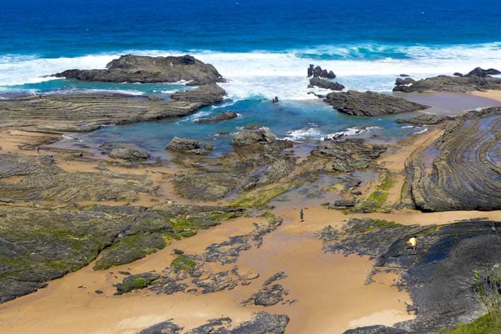 A view of a rocky beach is presented, featuring a mix of sand and stone formations. Gentle waves crash against the rocks, while various pools of water reflect the sky. The landscape is complemented by patches of green vegetation in the foreground.