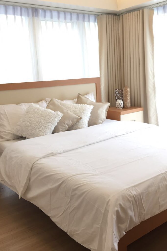 A bright bedroom features a spacious bed adorned with soft white linens and decorative pillows. Natural light flows through large windows, complemented by sheer curtains. A bedside table and decorative vases add a subtle touch to the clean and modern design.