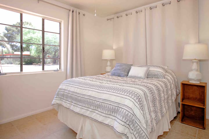 საძინებელში არის გრძელი ორმაგი საწოლი, ელეგანტური გეომეტრიული ლამპები, კარადის სარკიანი კარები და სახლის წინა მხარის ხედი.