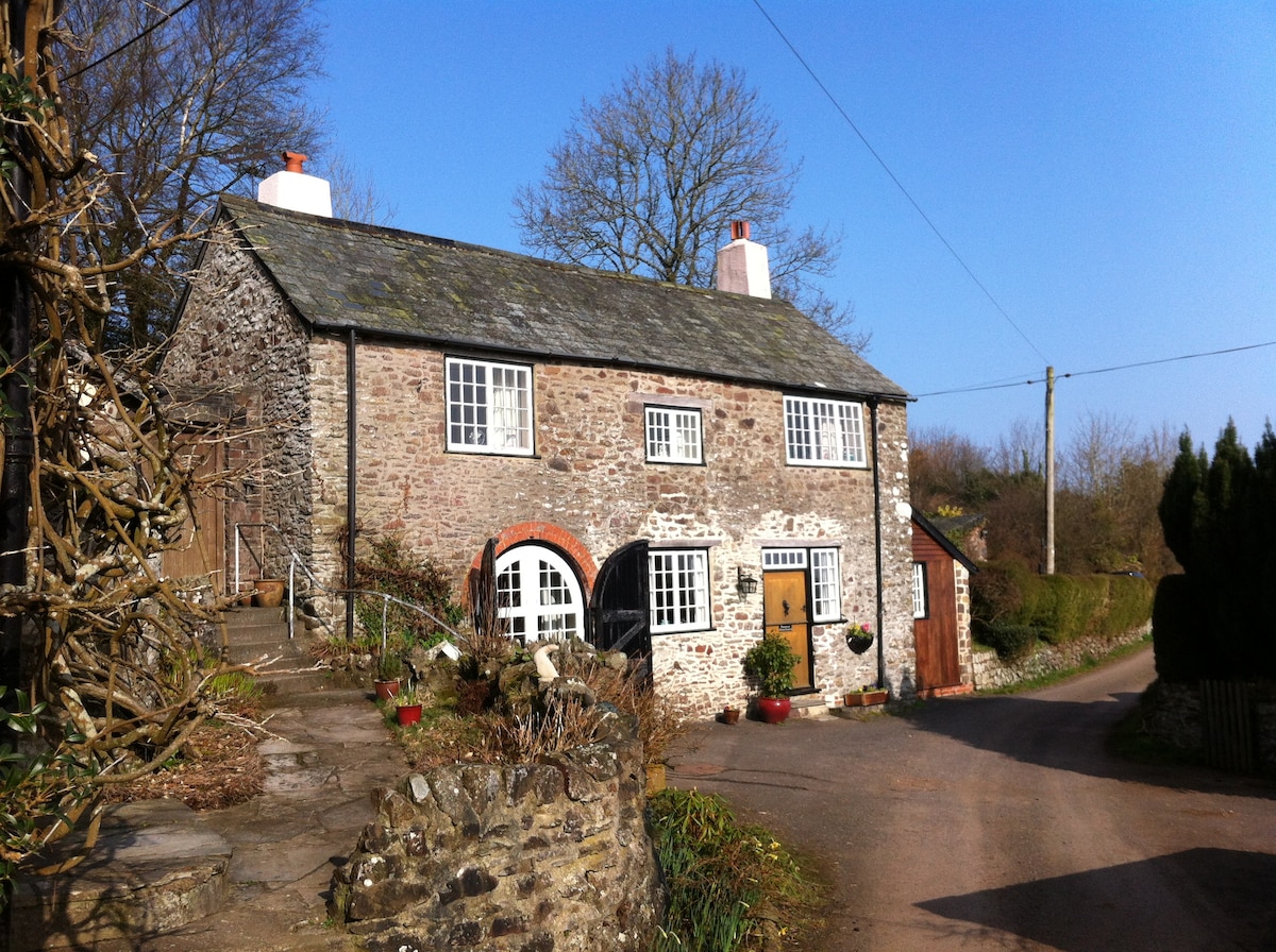 A traditional stone cottage is set against a clear blue sky, showcasing a welcoming exterior with white windows and red brick accents. The entrance features arched doorways, and flower pots line the pathway, adding a touch of greenery to the rustic landscape.