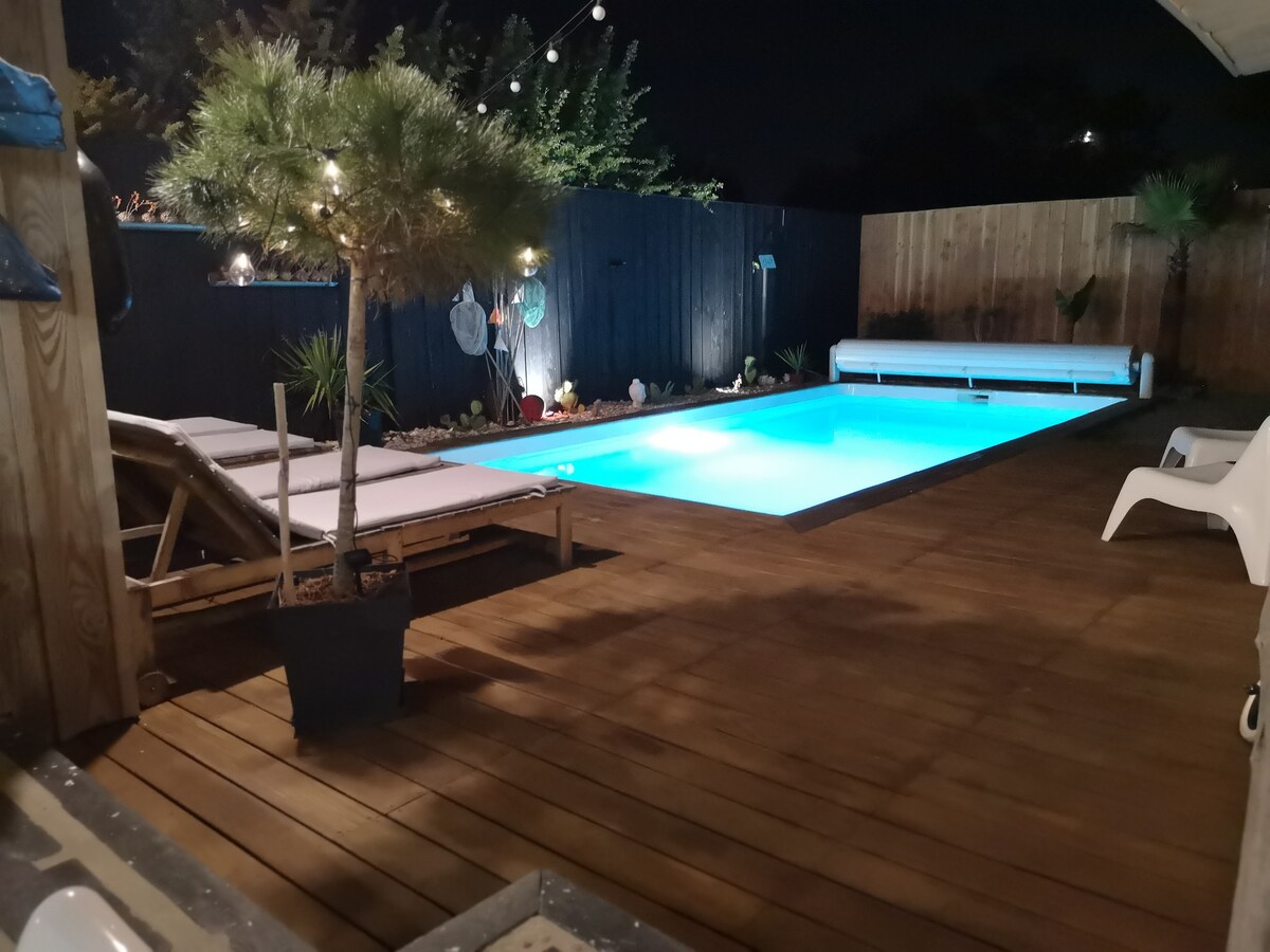 A serene evening scene features a heated swimming pool illuminated in soft blue light, surrounded by wooden decking. Two lounge chairs are positioned nearby, and potted plants add greenery to the area. Fairy lights gently illuminate the surrounding wooden fence.