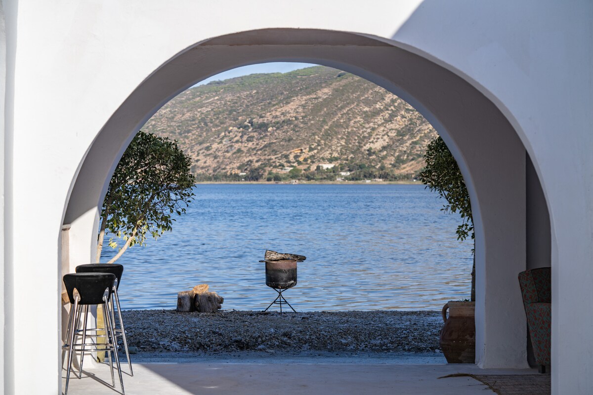 An archway frames a view of calm waters, with a grill positioned on the shoreline. Two small trees are visible on either side, and the gentle silhouette of hills serves as a backdrop, enhancing the peaceful setting.