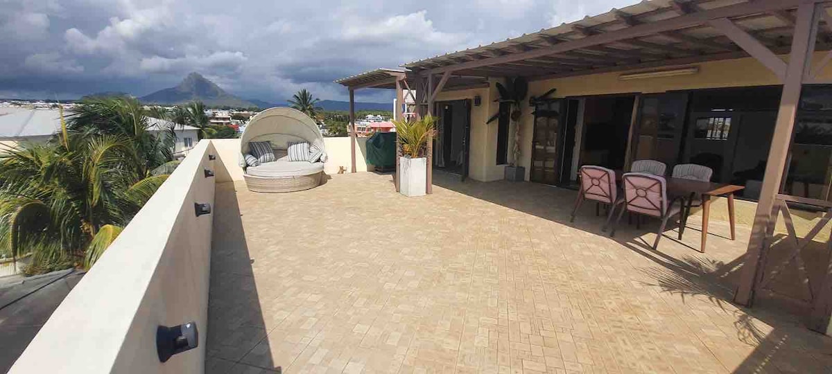 A spacious patio is shown, featuring a circular lounge bed and a dining table with chairs. Potted plants add greenery, while mountains are visible in the background. The area is covered by a pergola, providing shade, and the flooring consists of light-colored tiles.