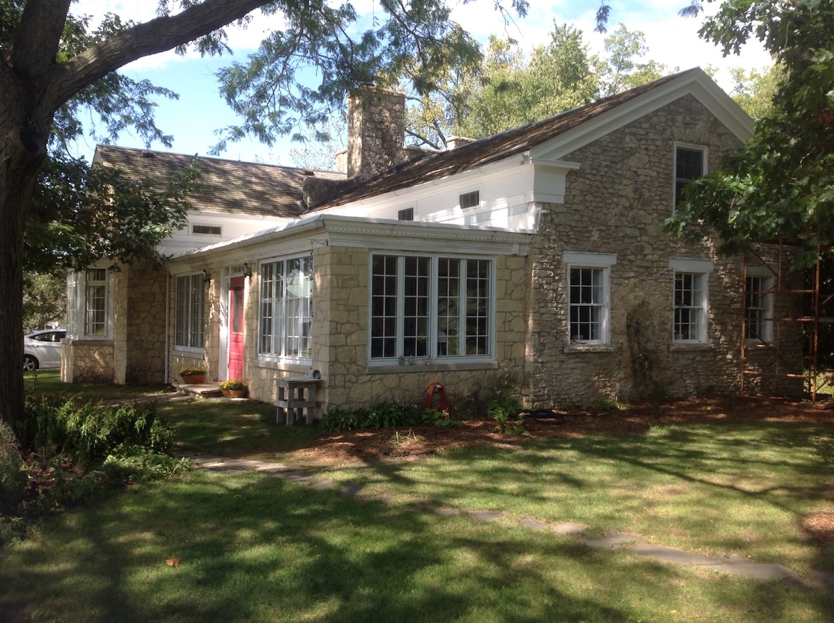 A historic limestone home is framed by lush greenery, showcasing large windows that invite natural light into the living spaces. The stone facade highlights the home's charm, while the well-maintained lawn offers a welcoming setting. A pathway leads toward the entrance, enhancing accessibility.