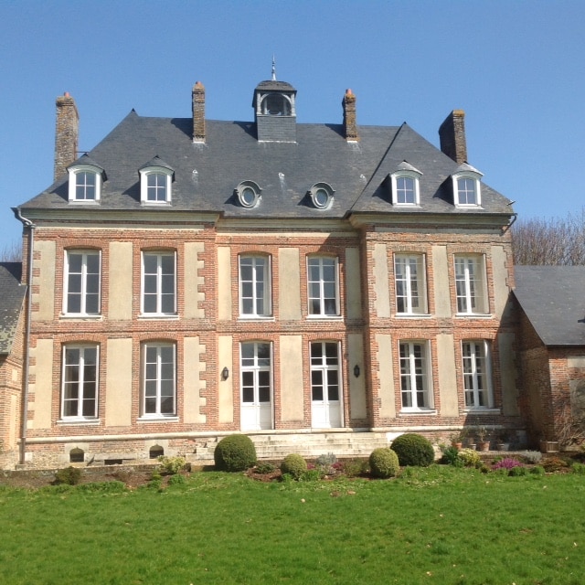 The elegant façade of the château showcases a blend of brick and stone construction, highlighted by multiple tall windows and decorative roof elements. A manicured lawn in front features trimmed bushes, contributing to the historic charm of the structure under a clear blue sky.