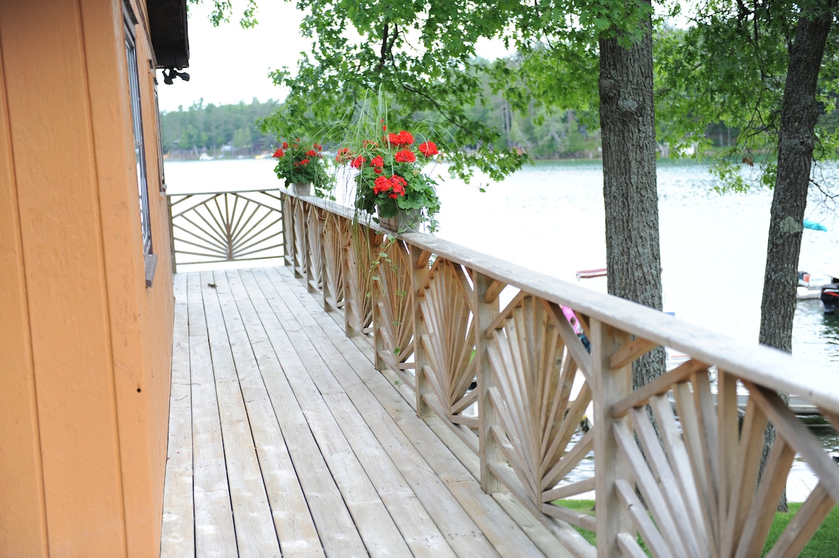 A wooden deck extends along the cabin's exterior, featuring a decorative railing with intricate designs. Hanging planters filled with vibrant red flowers provide a pop of color against the natural backdrop of trees and the lake, enhancing the outdoor space.