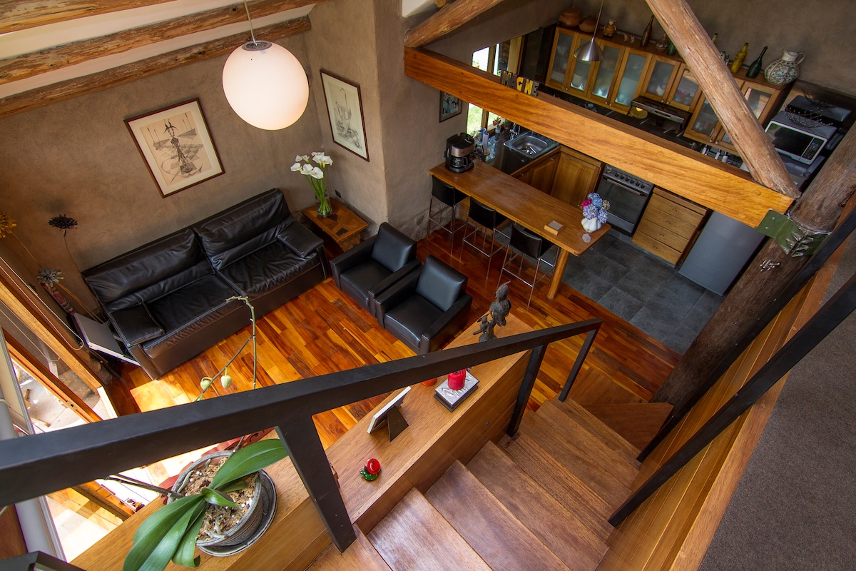 A view from above captures the spacious living area featuring hardwood floors and exposed wooden beams. A dark leather sofa and two armchairs are arranged around a low table. The open kitchen showcases a wooden island, blending with the overall rustic charm of the space.