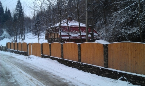 A wooden fence line runs alongside a snowy road, offering a glimpse of a chalet with a red roof nestled among trees. The landscape is blanketed in snow, enhancing the serene, quiet setting of the area. Power lines are visible in the background.