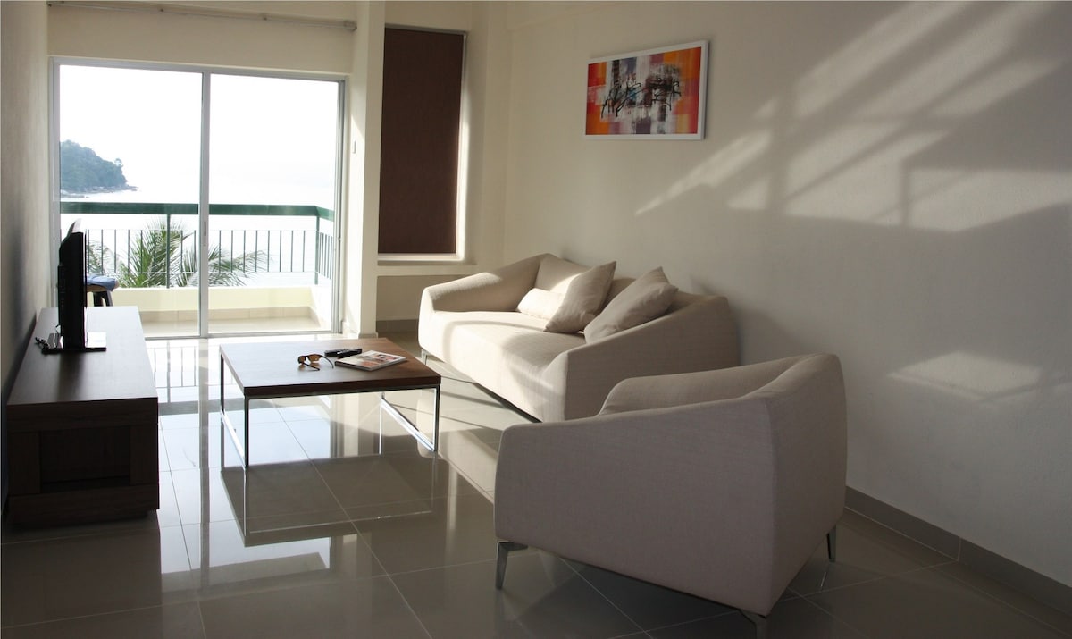 A bright living area features a comfortable sofa and armchair facing a coffee table. Natural light floods in through large glass doors, revealing a view of the balcony and the sea beyond. A piece of abstract wall art adds a splash of color.