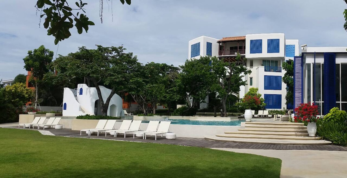 A serene pool area is surrounded by lush greenery and modern architecture. Sun loungers line the poolside, offering relaxation under the open sky. The vibrant building features blue accents and is complemented by well-tended gardens, creating a refreshing outdoor environment.