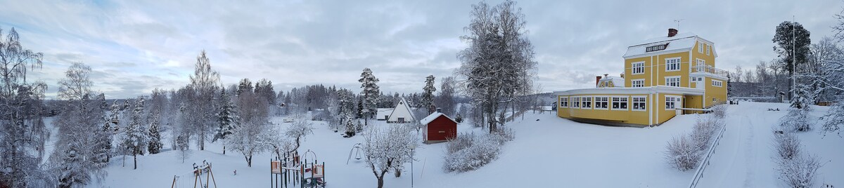 A panoramic view captures a winter landscape featuring a snow-covered playground with colorful equipment. A large yellow building stands prominently, surrounded by white-dusted trees and quaint structures, creating a serene and tranquil environment.