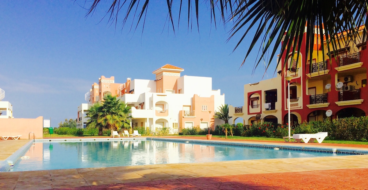 The image shows a serene swimming pool surrounded by landscaped gardens and palm trees. Multiple buildings with a mix of pastel colors can be seen in the background. Sun loungers are placed along the pool's edge, highlighting the inviting outdoor space.