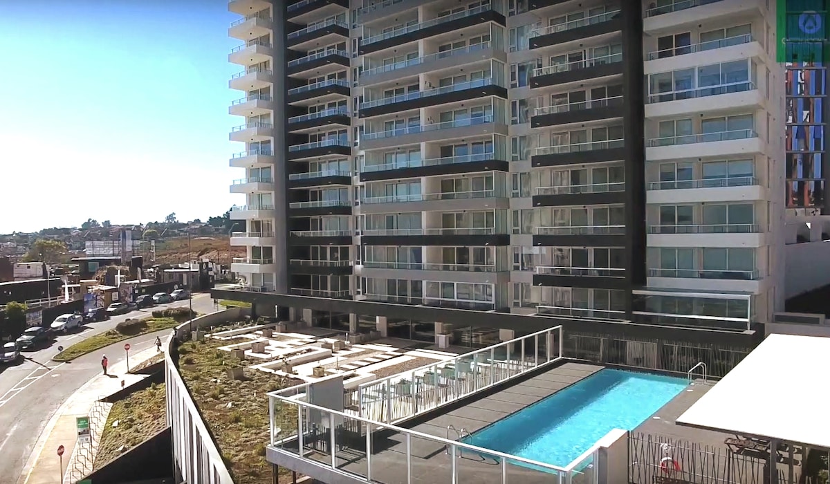 The image displays a modern residential building with multiple floors, featuring a pool area below. The pool is surrounded by a patio with lounge chairs, and the surroundings include greenery and pathways. Clear skies add to the inviting atmosphere of the community space.
