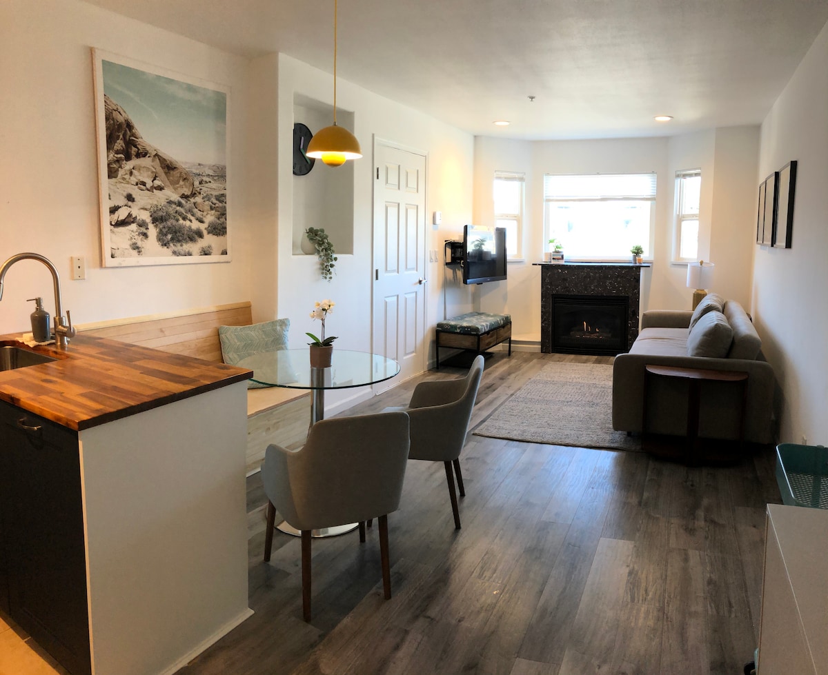 An open living space features a combination of a dining area with a glass table and plush chairs, alongside a cozy seating area. Natural light streams through multiple windows, illuminating the gas fireplace and complementing the calming neutral tones of the decor.