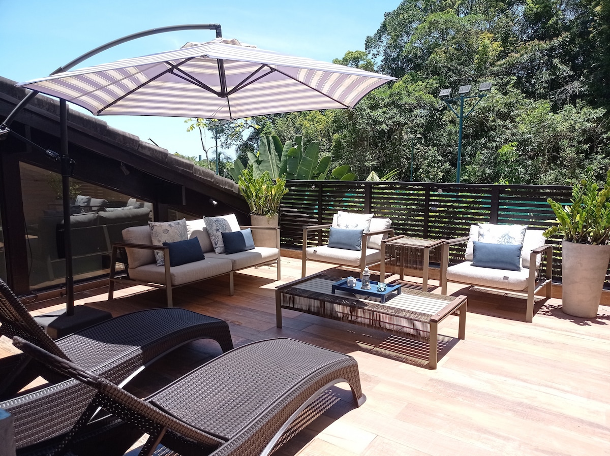 A spacious outdoor terrace features comfortable lounge chairs and a seating area with a coffee table, all set beneath a striped umbrella. Lush greenery is visible in the background, enhancing the tranquil atmosphere of this private space.