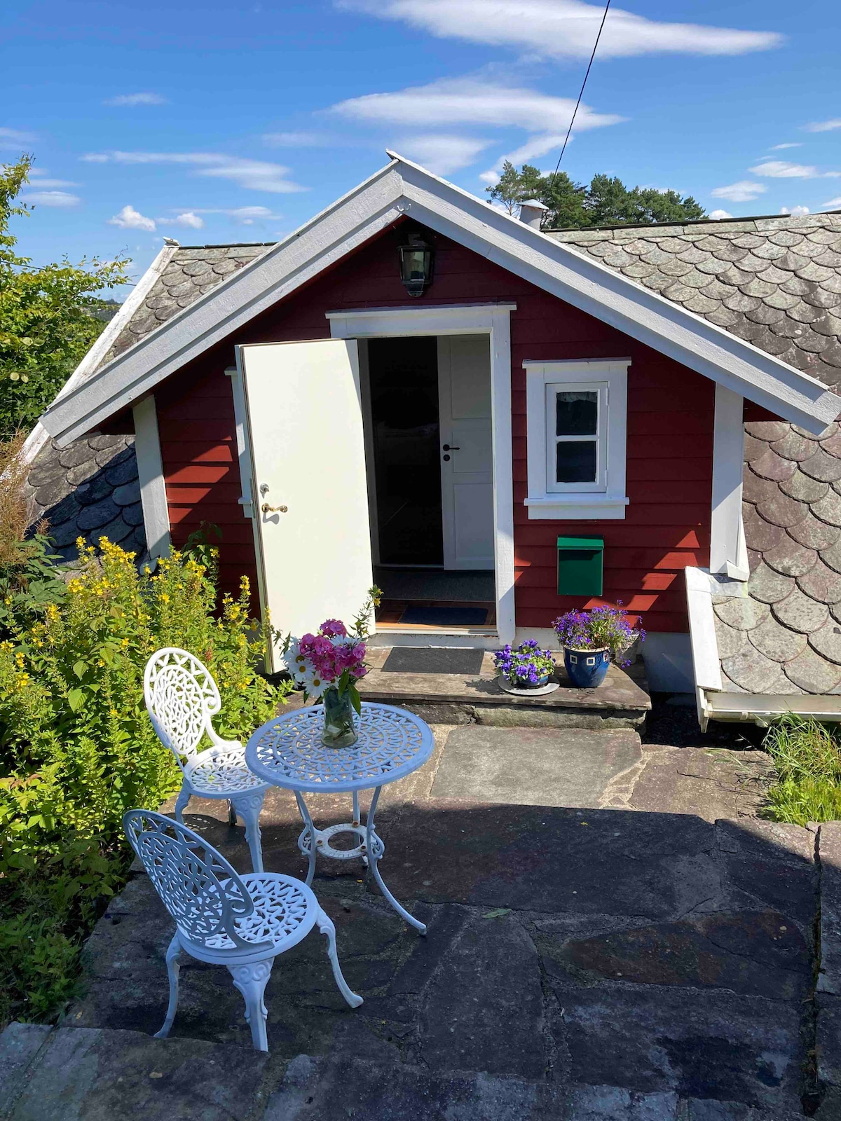 A charming entrance is framed by a red wooden exterior and a sloped roof. A small table with two white chairs is set on a stone path, accompanied by a vase of flowers. Bright greenery surrounds the area, enhancing the serene environment.