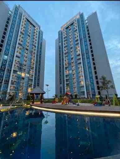 Two modern high-rise buildings are positioned beside a reflective infinity pool. Lush landscaping is visible around the pool, with seating areas and a children's play area. The evening sky provides a serene backdrop, enhancing the inviting outdoor space.