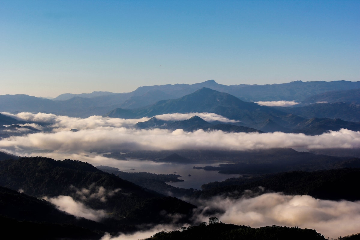 A panoramic view captures rolling mountains blanketed in soft clouds, with a serene lake visible below. The peaks rise majestically against a clear blue sky, creating a tranquil scene that showcases the natural beauty of the mountainous region.