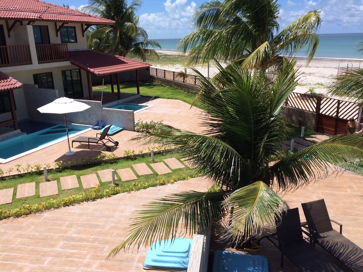 A sunlit outdoor area features a private pool surrounded by lush greenery and sun loungers. An umbrella provides shade for relaxation, while a pathway leads towards the beach. The serene sea can be seen in the background, contrasting with the vibrant blue sky.