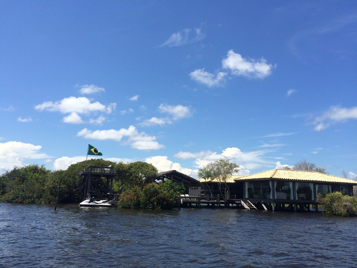 A striking view of the house, situated on the water's edge, is highlighted by the Brazilian flag waving in the breeze. Surrounding greenery adds to the serene ambiance, while the clear blue sky above complements the tranquil setting.