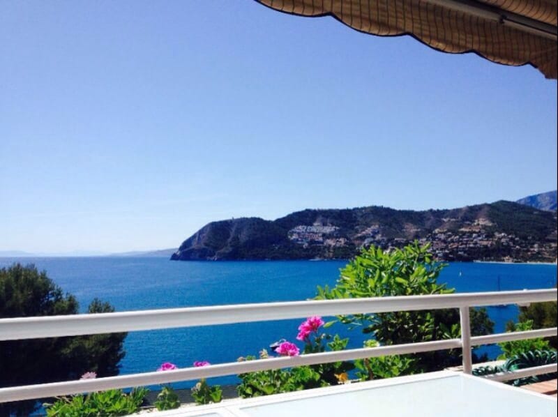 A panoramic view of the bay is presented from the terrace, showcasing vibrant blue waters and lush green vegetation. The coastline features gently sloping hills, and distant hills are visible under a clear blue sky, creating a serene backdrop.