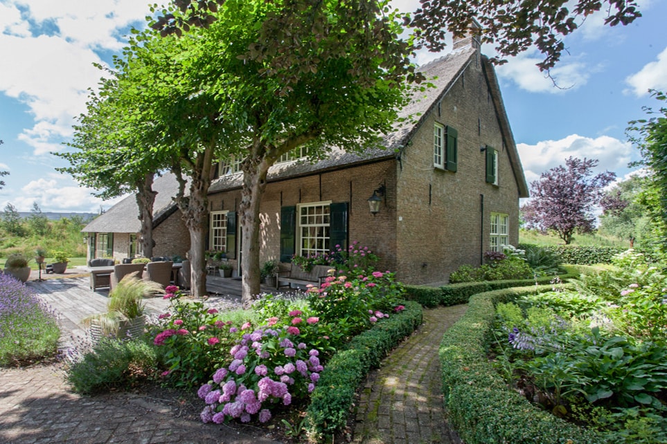 A classic restored farmhouse is surrounded by vibrant gardens filled with colorful flowers, including pink hydrangeas. The inviting outdoor space features a pathway leading to a spacious terrace adorned with seating areas, offering a peaceful view of the greenery.