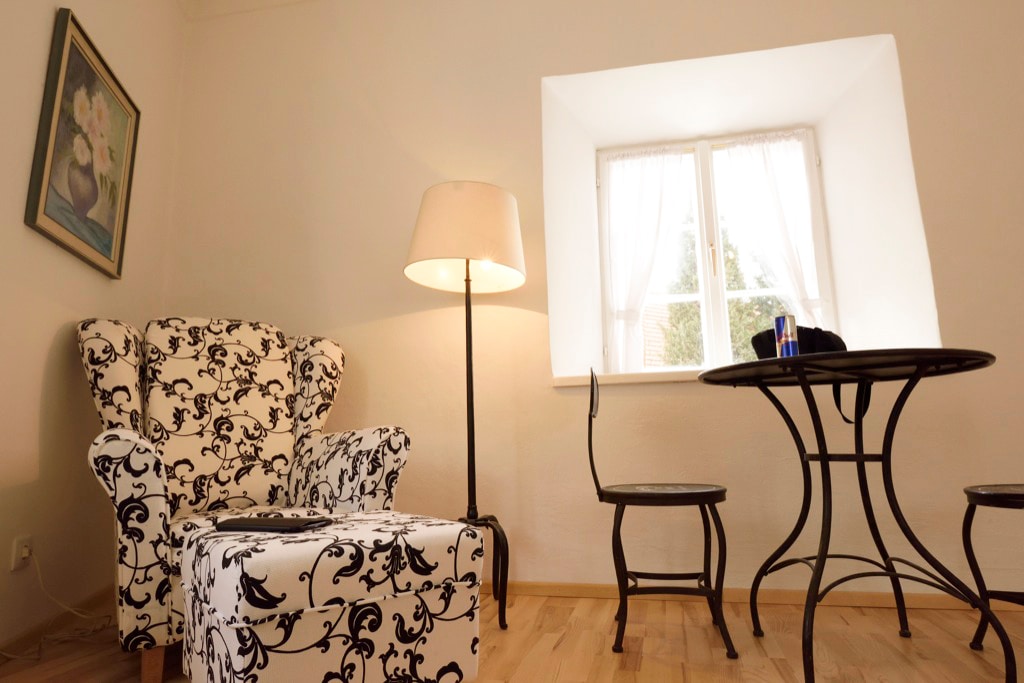 A seating area features a patterned armchair, a small round table, and a chair. Natural light enters through a window with sheer curtains. A floor lamp provides additional illumination, while a floral artwork adorns the wall.