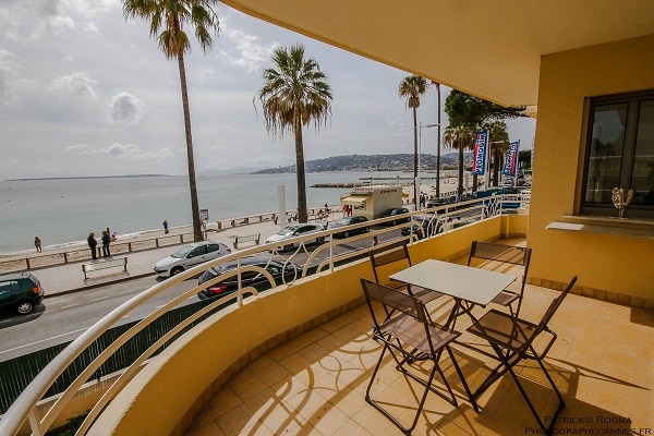 A large balcony offers views of the sea and nearby beach, framed by palm trees. A small dining table and two chairs are positioned on the tiled surface. The scene captures the relaxed atmosphere of the waterfront and nearby promenade.