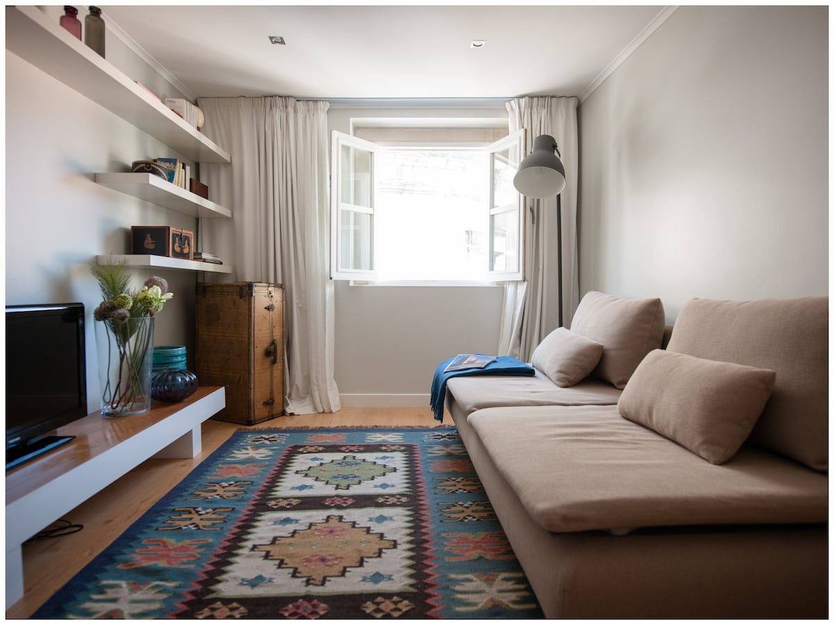 A light-filled living area features a comfortable sofa and decorative shelves with curated items. A large window opens to reveal flowing curtains, allowing natural light to brighten the space. A colorful patterned rug adds warmth to the hardwood floor.