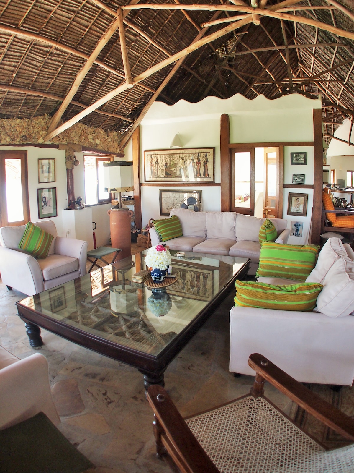 A spacious living area features a glass coffee table at its center, surrounded by comfortable seating, including a sectional sofa and vintage chairs. Natural materials are highlighted in the architecture, with a thatched ceiling and decorative wall art adding character.