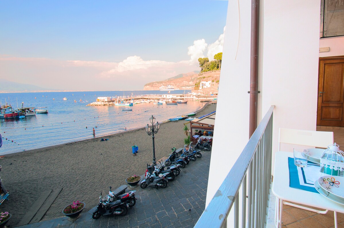 A view from the apartment's balcony showcases the sandy beach of Marina Grande, with the sea gently lapping at the shore. Fishing boats are anchored in the water, and a row of scooters is parked along the promenade. Tables are set for outdoor dining, allowing for a pleasant seaside experience.