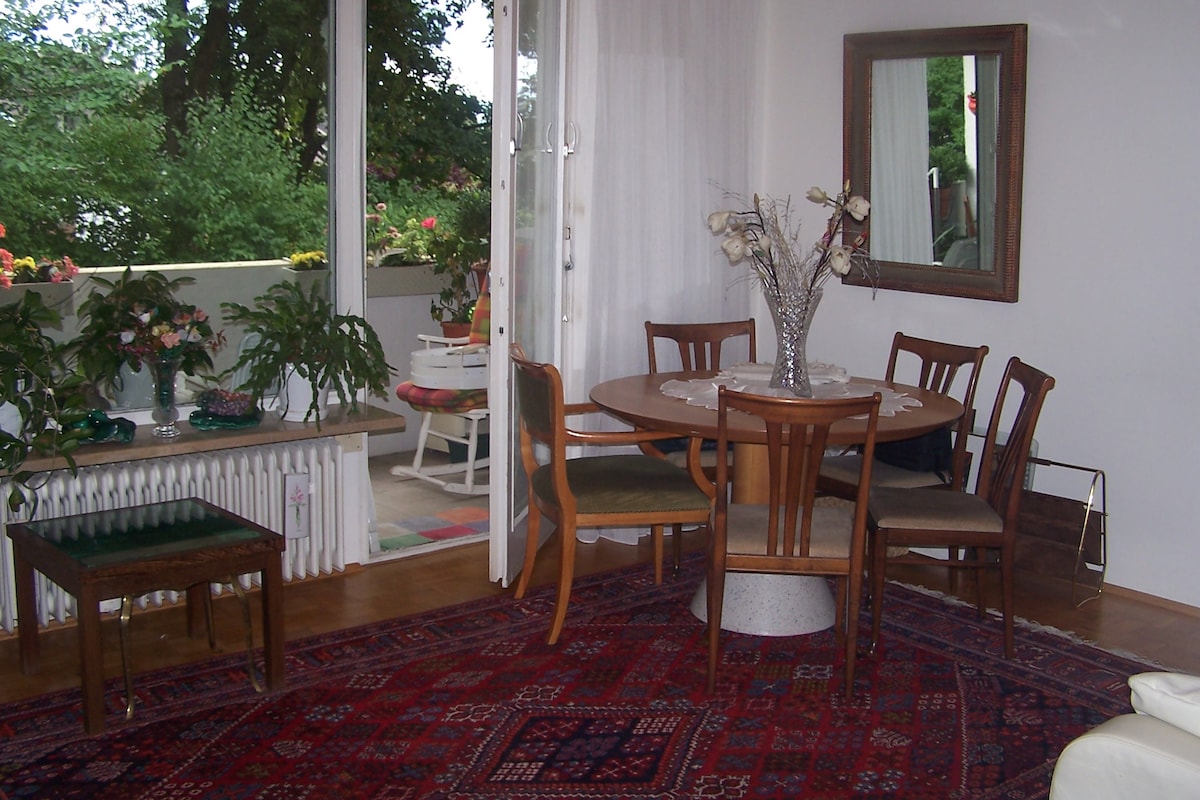 A dining area is featured with a round wooden table surrounded by six chairs, set on a patterned red rug. A large mirror is mounted on the wall, reflecting the natural light from a nearby balcony, which showcases greenery and floral arrangements.