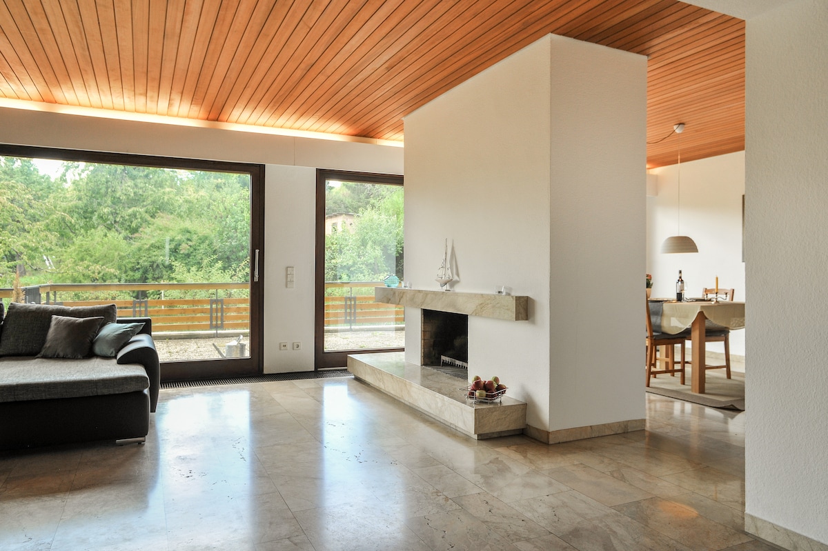 A bright living area features a marble floor with a modern, minimalist design. Large glass doors provide natural light and views of the greenery outside. A cozy seating area surrounds a stone fireplace, with a dining space visible in the background.