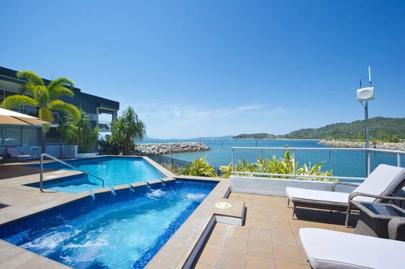 A tranquil pool area features a rectangular pool adjacent to a spa, with views of the harbor and surrounding greenery. Sun loungers are positioned nearby, inviting relaxation under a clear blue sky.