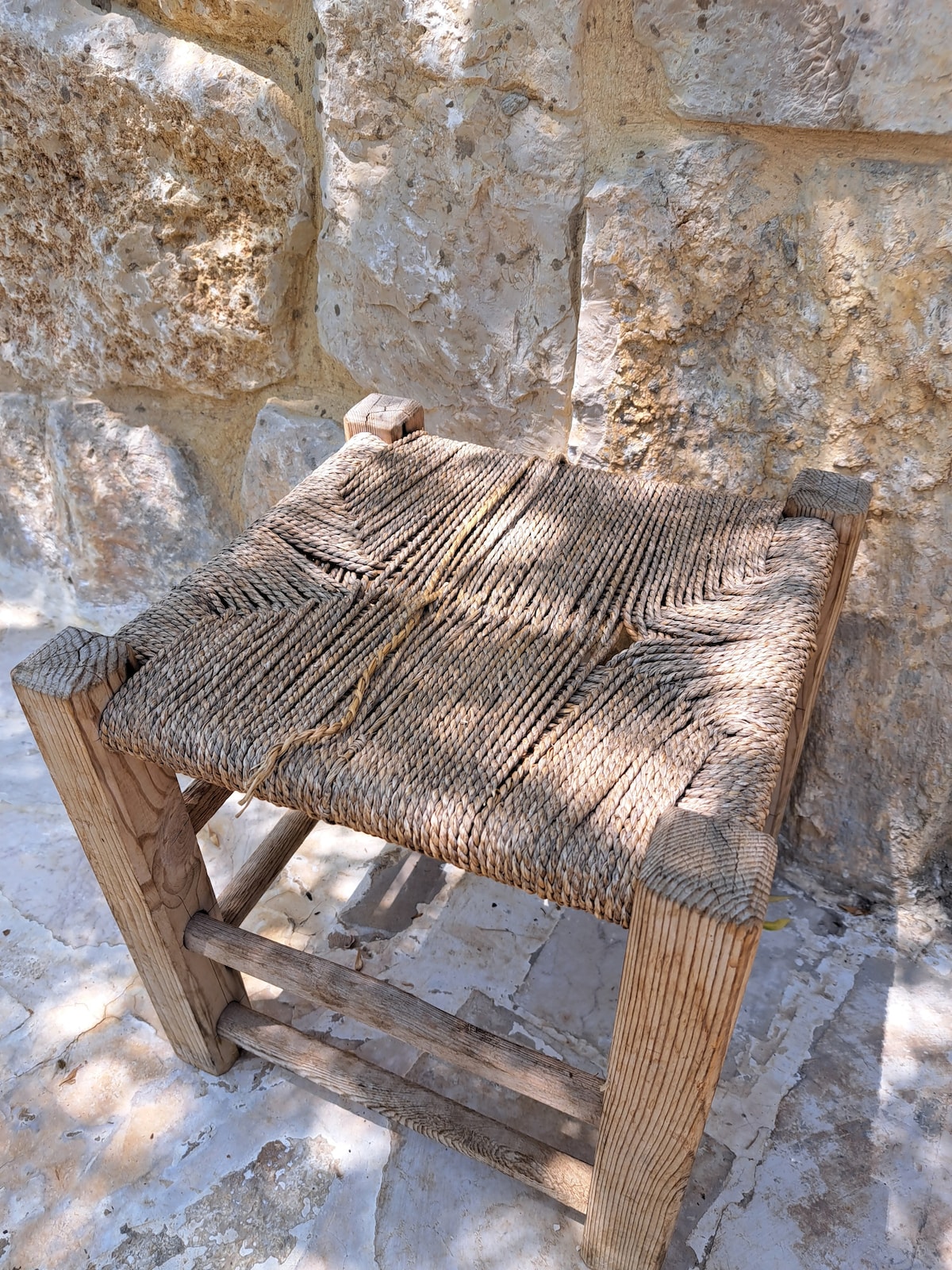A rustic wooden stool features a woven seat made from natural fibers, resting against a textured stone wall. The stool's frame is sturdy, highlighting the craftsmanship in its simple design. Natural light subtly enhances its organic appearance.