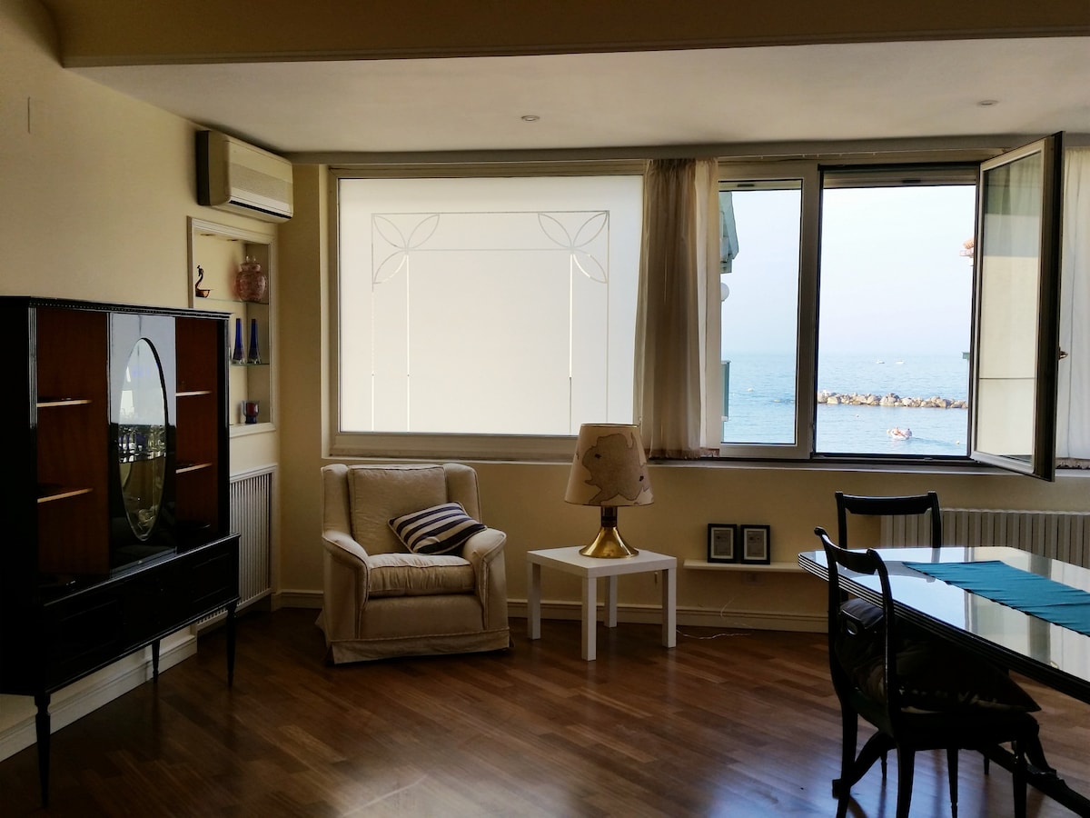 A bright living area features large windows offering a view of the sea. A comfortable armchair is positioned beside a small table under a lamp. Dark wooden furniture adds contrast to the light walls, creating a calm and inviting space.