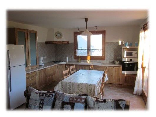 A spacious kitchen area is equipped with various wooden cabinets and modern appliances, including a refrigerator, oven, and stovetop. A large table with a patterned tablecloth is surrounded by wooden chairs. Natural light filters through the large window, enhancing the inviting atmosphere.