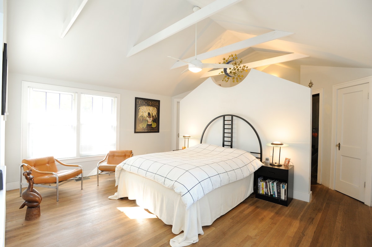 The spacious master suite features a queen bed adorned with a white coverlet, flanked by two modern bedside lamps. Two leather chairs provide a seating area, while large windows offer ample natural light and views of the surrounding neighborhood.