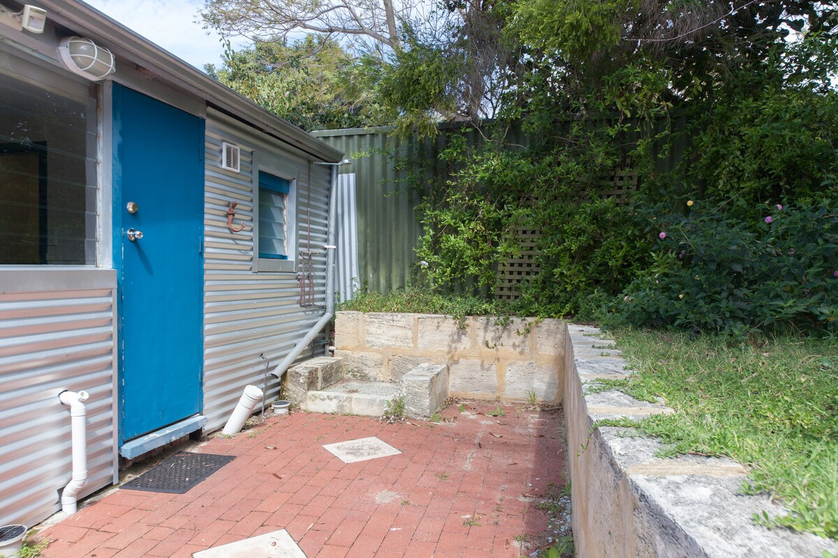 An outdoor studio is presented with a vibrant blue door set against a backdrop of green foliage. A paved area with stone steps and a small grassy patch is visible, providing a calm and inviting space for relaxation.